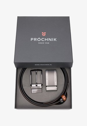 Brązowy skórzany pasek z dwoma klamrami: klasyczną czarną w stylu, oraz nowoczesną srebrną z rytowanymi detalami, umieszczony w czarnym pudełku.