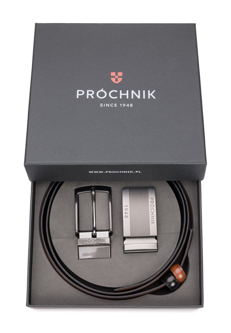 Brązowy skórzany pasek z dwoma klamrami: klasyczną czarną w stylu, oraz nowoczesną srebrną z rytowanymi detalami, umieszczony w czarnym pudełku.