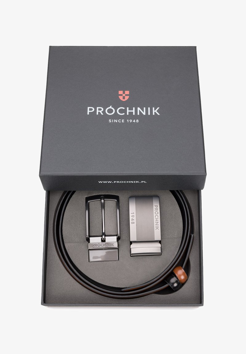 Brązowy skórzany pasek z dwoma klamrami: klasyczną czarną w stylu, oraz nowoczesną srebrną z rytowanymi detalami, umieszczony w czarnym pudełku.