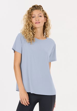 Vrouw met krullend blond haar draagt een lichtblauw T-shirt met korte mouwen en zwarte broek, staand tegen een effen witte achtergrond.