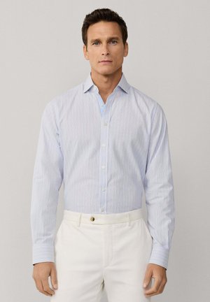 Hombre con camisa de botones a rayas azul claro, metida dentro de pantalones blancos, de pie frente a un fondo gris claro liso.