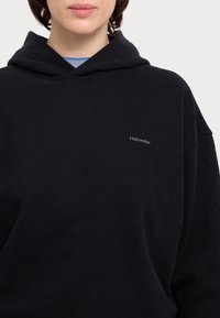 Felpa nera con cappuccio, realizzata in morbido tessuto misto cotone, con tasca frontale, cappuccio regolabile con cordini e logo sottile "Holzweiler" sul petto.