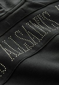 Tessuto nero con una texture liscia presenta la parola "WASSUP" in strass argentati, disposti in un design curvo lungo la cucitura.
