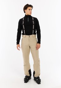 Beige skibroek met zwarte accenten, verstelbare bretels, twee voorzakken en een slank ontwerp, gecombineerd met een zwart zip-hals shirt en skischoenen.