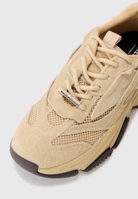 Zapatilla beige que cuenta con materiales de ante y malla, puntera redonda, cierre con cordones y suela de goma con acentos en marrón oscuro.