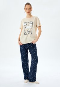 Beige Baumwoll-T-Shirt mit "NAP"-Grafik, kombiniert mit dunkelblauen Schlafanzughosen mit einem weißen himmlischen Muster. Weicher, lockerer Schnitt, kurze Ärmel.