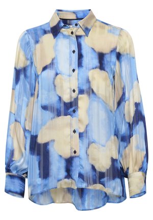 Persoon met kort haar die een blauw en beige gebloemde blouse draagt, lichtblauwe hoge taille broek en zwarte pluizige sandalen, staand met de rug naar ons toe.