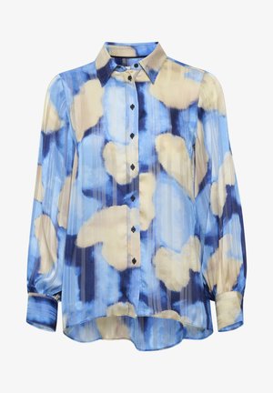 Persoon met kort haar die een blauw en beige gebloemde blouse draagt, lichtblauwe hoge taille broek en zwarte pluizige sandalen, staand met de rug naar ons toe.