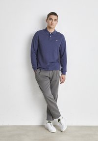 Langärmliges lila Polo-Shirt mit Kragen, kombiniert mit grauen Hosen und weißen Sneakern. Das Design ist einfach gehalten mit einem Logo-Akzent auf der Brust.