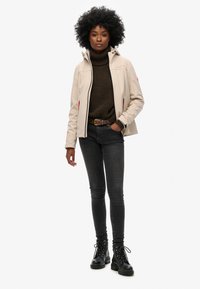 Beige jas met hoge kraag, rits aan de voorkant en rode accenten, gecombineerd met een bruine coltrui en zwarte skinny jeans. Zwarte veterboots.
