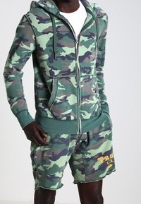 Sudadera con cremallera de camuflaje con bolsillos y detalles en verde, combinada con pantalones cortos de camuflaje a juego con un dobladillo deshilachado y detalle de texto en naranja.