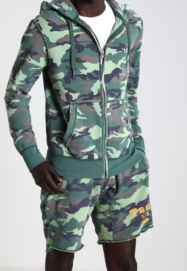 Sudadera con cremallera de camuflaje con bolsillos y detalles en verde, combinada con pantalones cortos de camuflaje a juego con un dobladillo deshilachado y detalle de texto en naranja.
