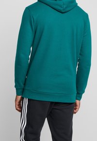 Person klädd i en turkos huvtröja och svarta byxor med vita sidoflikar, sedd bakifrån mot en enfärgad bakgrund.