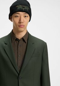 Grön skräddarsydd blazer över en brun knäppt skjorta, i kombination med en svart stickad mössa med grön "HUGO" brodering.
