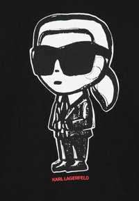 Crna grafička majica s bijelom ilustracijom figure u naočalama, odjevenoj u odijelo, s natpisom "KARL LAGERFELD" ispisanim crveno ispod.