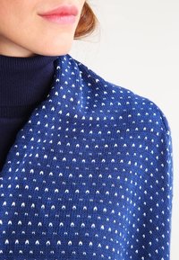 Kvinna iklädd en blå mönstrad sjal över en mörkblå polotröja, som visar läppar och haka mot en vit bakgrund.