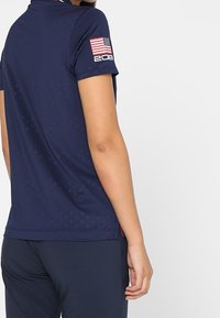 Mörkblå atletisk polo med krage, korta ärmar och en amerikansk flagga på höger ärm. Har subtila texturer och designinslag.