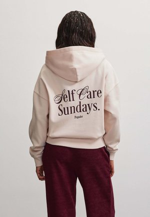 Persoon met gevlochten haar draagt een lichte hoodie met tekst "Self Care Sundays" op de rug en donkerrode broek, staand met de rug naar voren.