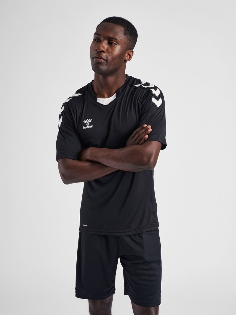 Mann mit verschränkten Armen, trägt ein schwarzes hummel Sporttrikot und Shorts vor einem einfarbigen grauen Hintergrund.