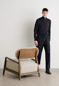Svart zip-up genser og svarte cargo-bukser båret av en person ved siden av en stol med treramme og en beige pute i sete og ryggstøtte.