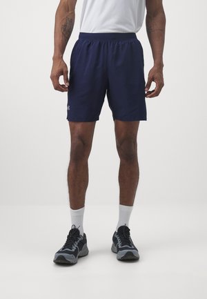 Mann trägt marineblaue Sportschuhe, weiße Socken und schwarze sowie graue Laufschuhe, steht vor einem weißen Hintergrund.