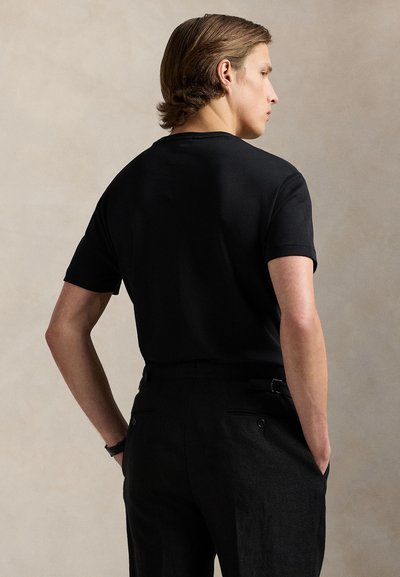 Ein junger Mann mit hellbraunen Haaren, der ein schwarzes kurzärmeliges Hemd und schwarze Hose trägt, steht mit den Händen in den Taschen seitlich vor einem beigen Hintergrund.