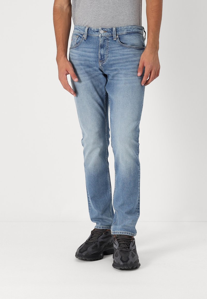 Muž v bleděmodrých vybledlých slim-fit džínách a černých sportovních botách, stojící proti čistému bílému pozadí.