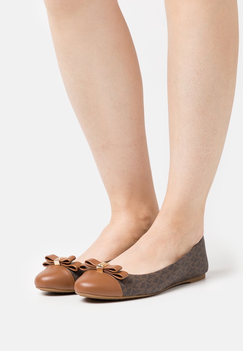 Chaussures plates marron et beige avec des nœuds sur les orteils portées sur des jambes nues sur un fond blanc uni.