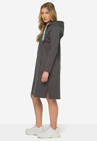Robe à capuche grise en matériau doux, dotée d'une poche avant, de fentes sur les côtés et d'un cordon contrastant vert. Assortie à des baskets blanches.