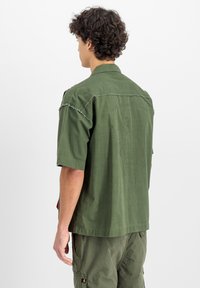 Camicia verde a maniche corte con vestibilità rilassata, cuciture delle spalle sfrangiate e un design grafico sul retro. Realizzata in leggero tessuto di cotone.