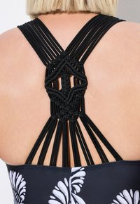 Černé plavky s designem křížových popruhů na zadní straně, s texturovaným makramé detailem uprostřed a květinovým vzorem na přední straně.