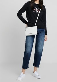 Czarna bluza z kapturem z logo "Calvin Klein Jeans", niebieskie dżinsy, biała torba na ramię i białe trampki. Prosty design, swobodny krój.