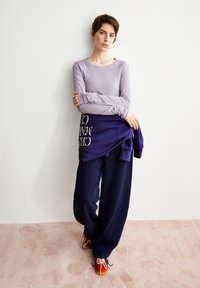 Junge Person mit kurzen Haaren steht vor einer weißen Wand und trägt ein hellviolettes Langarmshirt, weite marineblaue Hosen und rote Sneaker.