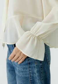 Hand resting near pocket wearing sheer, cream-colored blouse with gathered bell sleeve and blue denim jeans.