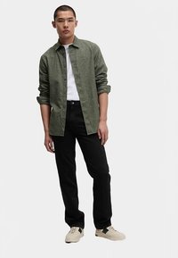 Grön texturerad skjorta, vit t-shirt, svarta jeans, cremefärgade sneakers med gröna detaljer. Modellen står med armarna längs sidorna mot en enfärgad bakgrund.