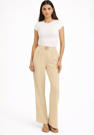 Femme portant un haut blanc ajusté à manches courtes, un pantalon large beige taille haute avec une ceinture décorative, et des sandales marron clair.