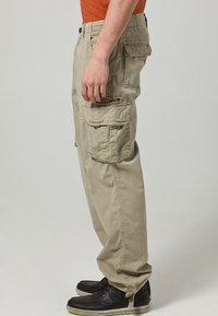 Beige cargo kalhoty s několika kapsami, elastickým pasem a volným střihem, vyrobené z měkké bavlněné směsi. Doplňují je černé tenisky.