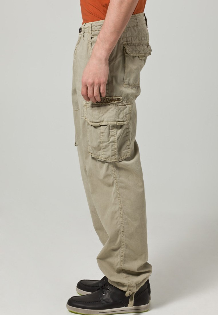 Beżowe spodnie cargo z wieloma kieszeniami, elastycznym pasem i luźnym krojem, wykonane z miękkiej mieszanki bawełny. Połączone z czarnymi sneakersami.
