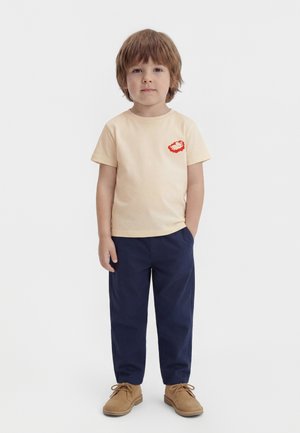 Fiatal fiú előre nézve, bézs színű pólót visel piros logóval, sötétkék nadrágot és világosbarna fűzős cipőt, egyszínű fehér háttér előtt.