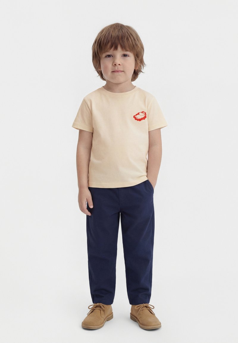 Νεαρός αγόρι στέκεται απέναντι φορώντας μπεζ μπλουζάκι με κόκκινο λογότυπο, ναυτικό μπλε παντελόνι και ταμπά δερμάτινα παπούτσια με κορδόνια, σε λευκό απλό φόντο.