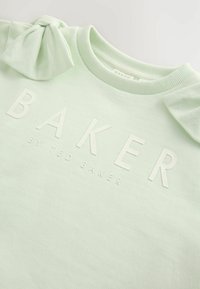 Gai zaļš džemperis ar gludu tekstūru, ar loku detaļu uz pleca un reljefu tekstu "BAKER by TED BAKER" baltā krāsā.