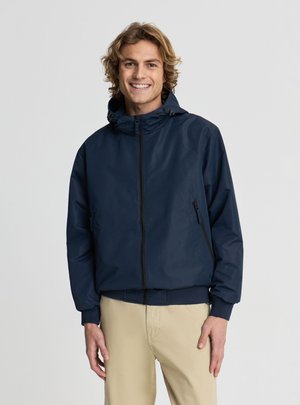 Uśmiechnięty młody mężczyzna z falującymi blond włosami w granatowej kurtce z kapturem i beżowych spodniach, stojący na jednolitym tle.