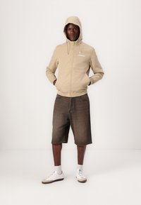 Ljust beige zip-jacka med huva, med en logotyp fram. Matchad med knälånga mörk denimshorts och vita sneakers med bruna sulor.