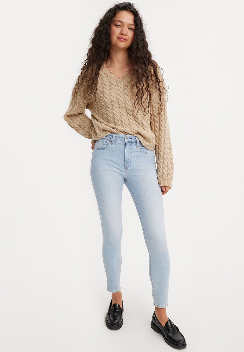 Pull en tricot beige avec un col en V et un motif en cable, associé à un jean skinny taille haute bleu clair et des mocassins noirs.