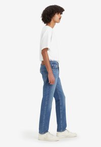 Jeans en denim bleu clair avec une coupe droite, taille mi-haute, cinq poches et une étiquette en cuir beige à l'arrière. Portés avec un t-shirt blanc.