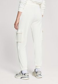 Białe spodnie cargo wykonane z miękkiego materiału, z bocznymi kieszeniami i elastycznymi mankietami przy kostkach, noszone z szarymi trampkami.