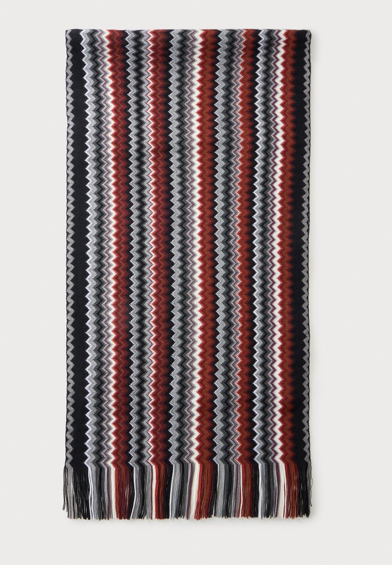 Écharpe tricotée avec des rayures zigzag verticales en noir, gris, blanc et couleur rouille, avec des franges aux deux extrémités.