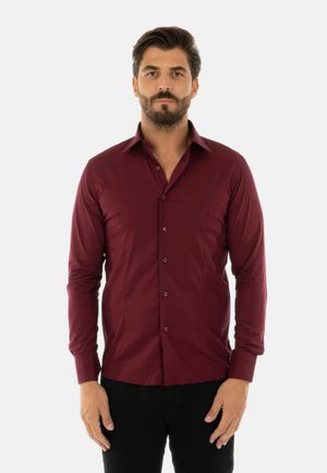 Uomo con capelli scuri e barba che indossa una camicia bordeaux aderente a maniche lunghe e pantaloni neri, in piedi davanti a uno sfondo neutro.