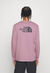 Tričko s dlouhými rukávy, světle fialové barvy, vyrobené z bavlny, se černým logem na zadní straně, kulatým výstřihem a volným střihem.