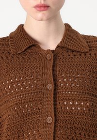 Cárdigan marrón tejido a mano con cuello de solapa, cierre de botones y detalles de tejido calado. Presenta una tela texturizada con un ligero brillo.